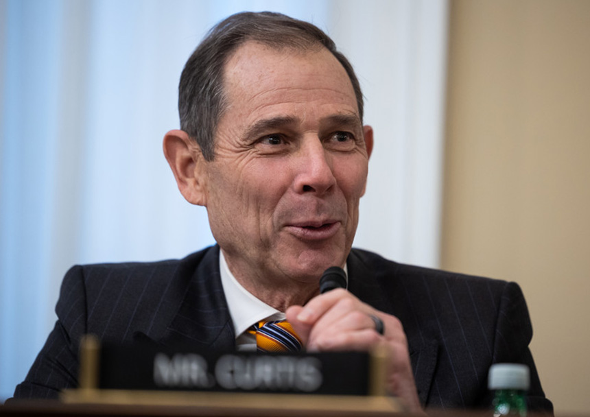 John Curtis holding a microphone while speaking from a dais.